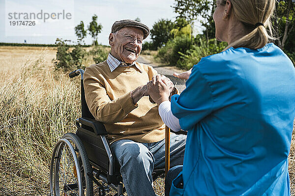 Glücklicher älterer Mann  der an einem sonnigen Tag im Rollstuhl sitzt und mit der Krankenschwester spricht