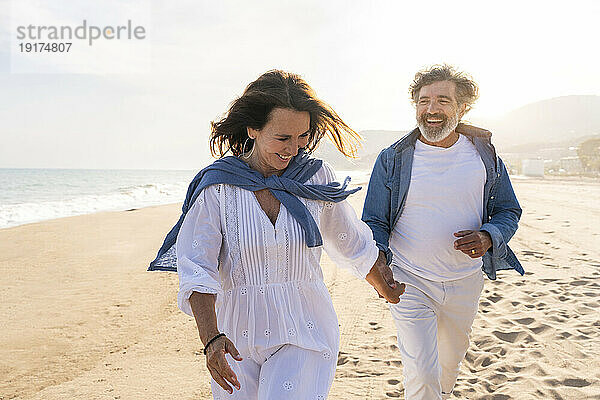 Fröhliche ältere Frau hält Händchen mit Mann und rennt am Strand