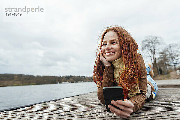 Lächelnde Frau mit Mobiltelefon liegt am Pier am See