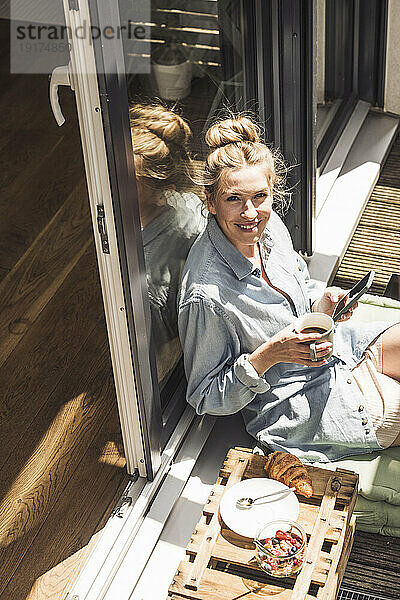 Porträt einer Frau  die sich mit einer Tasse und einem Smartphone in der Hand auf dem Balkon entspannt