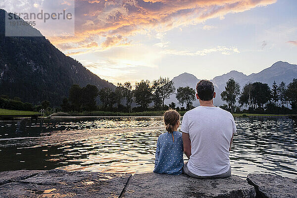 Vater und Tochter sitzen auf einem Felsen und bewundern den See bei Sonnenuntergang