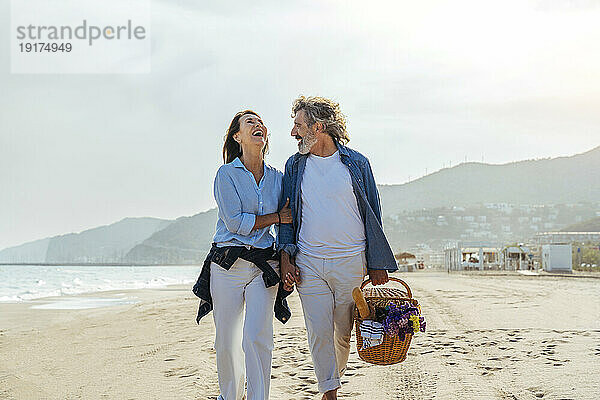Fröhliche Frau geht mit Mann mit Korb am Strand spazieren