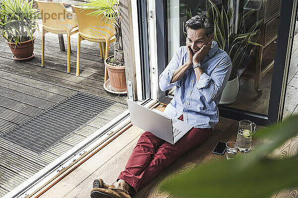 Portrait of happy man using laptop by open balcony