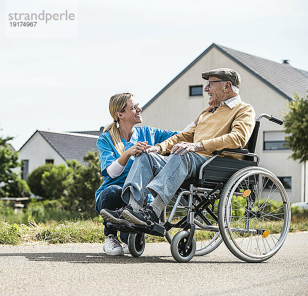 Lächelnde Krankenschwester im Gespräch mit einem älteren Mann  der an einem sonnigen Tag im Rollstuhl sitzt