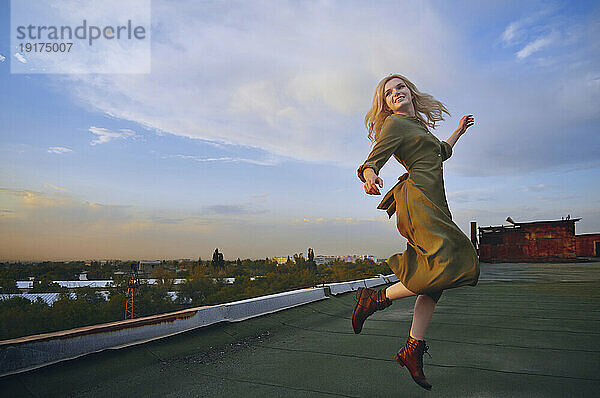 Cheerful blond woman dancing on rooftop under cloudy sky
