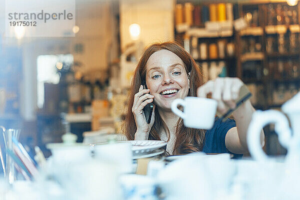 Lächelnde rothaarige Frau  die mit dem Smartphone telefoniert und im Laden Geschirr kauft