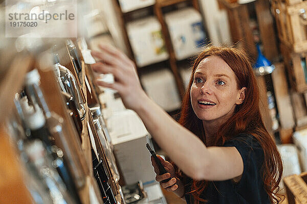 Lächelnde rothaarige Frau mit Smartphone greift im Geschäft nach Waren