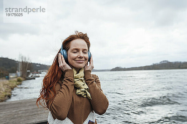 Lächelnde Frau hört Musik mit geschlossenen Augen am See