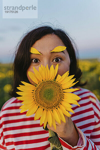 Reife Frau mit Blütenblättern auf den Augenbrauen  die den Mund mit einer Sonnenblume im Feld bedecken