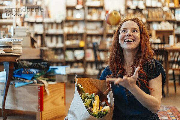 Fröhliche Frau gestikuliert und hält eine Tüte mit Lebensmitteln im Laden