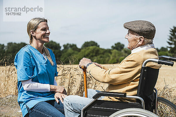 Lächelnde Krankenschwester im Gespräch mit einem älteren Mann  der an einem sonnigen Tag im Rollstuhl sitzt