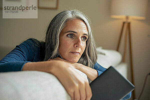 Thoughtful mature woman sitting on sofa with tablet PC