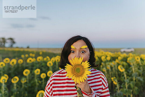 Reife Frau mit Blütenblättern auf den Augenbrauen  die den Mund mit Sonnenblumen bedecken