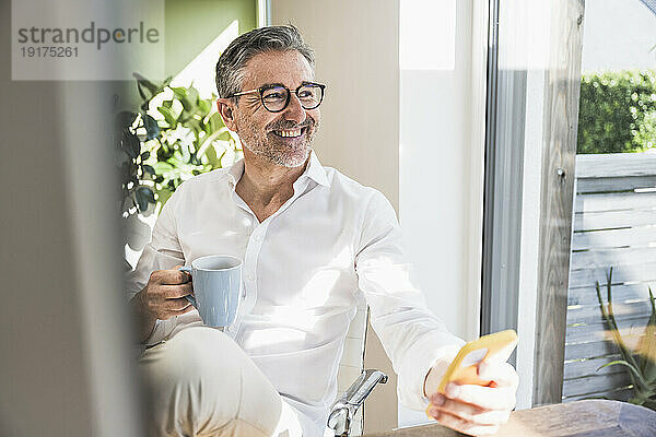 Nachdenklicher Freiberufler mit Smartphone und Kaffeetasse im Homeoffice