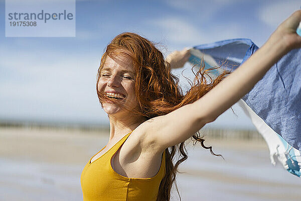 Cheerful woman with tousled hair playing with scarf on beach holiday