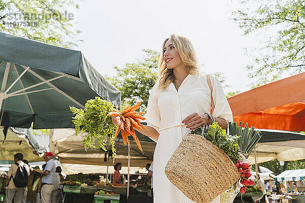 Lächelnde Frau hält einen Haufen Karotten und eine Tüte Lebensmittel auf dem Markt