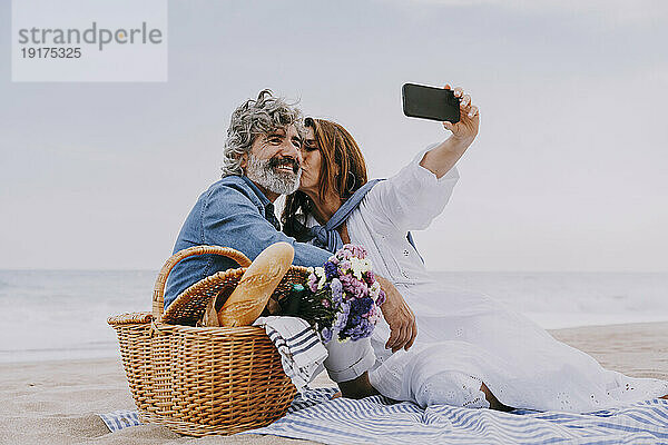 Glückliche Seniorin küsst Mann und macht Selfie mit Smartphone am Strand