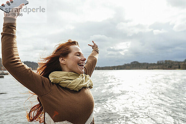 Glückliche Frau mit Mobiltelefon am See unter bewölktem Himmel