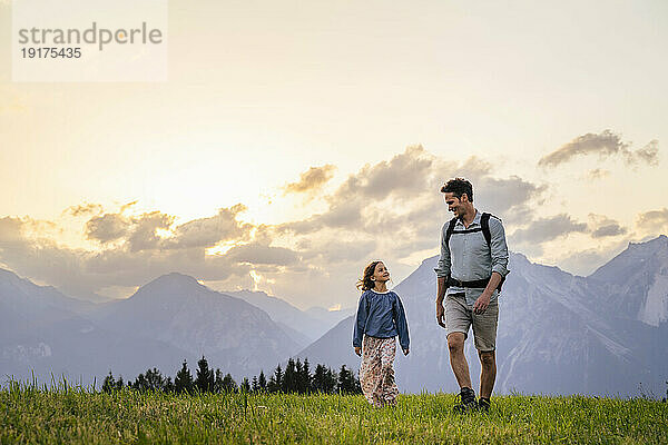 Father and daughter walking on grass in front of mountain range at sunset
