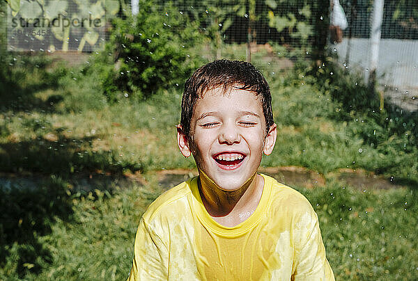 Fröhlicher Junge  der an einem sonnigen Tag im Hinterhof mit Wasser spielt