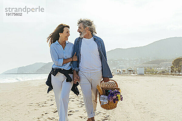 Glückliche Frau geht mit Mann mit Korb am Strand spazieren