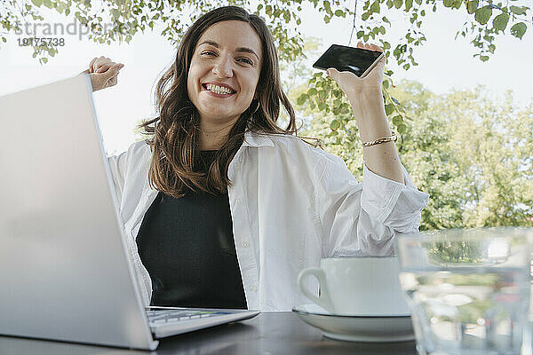 Glückliche Geschäftsfrau sitzt mit Laptop und hält Smartphone am Tisch im Garten