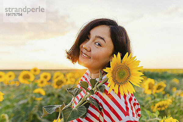 Reife lächelnde Frau mit Sonnenblume bei Sonnenuntergang