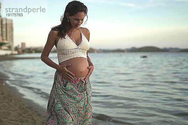 Schwangere Frau mit den Händen auf dem Bauch steht am Strand