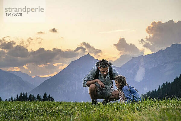 Father and daughter squatting on grass in front of mountains