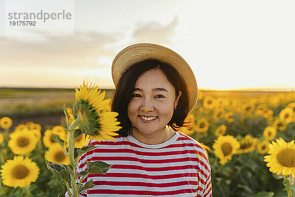 Lächelnde Frau mit Hut hält Sonnenblume im Feld bei Sonnenuntergang