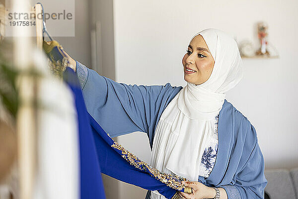 Fröhlicher junger Modedesigner mit Hijab und Blick auf das blaue Hochzeitskleid