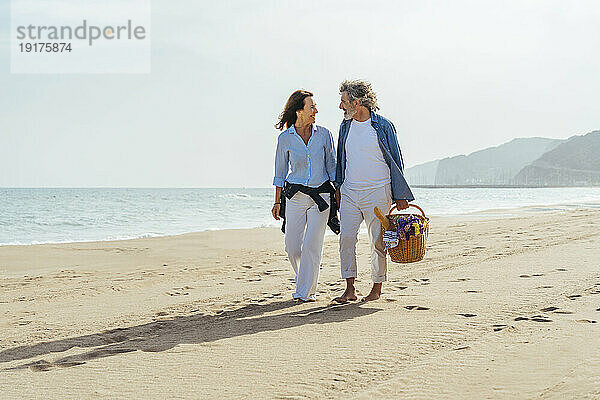 Glückliches älteres Paar hält Händchen und geht mit Korb am Strand spazieren