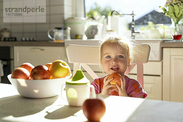 Happy girl holding apple fruit sitting at dining table in kitchen