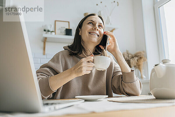 Lächelnder Freiberufler  der mit dem Smartphone spricht und eine Teetasse in der Küche hält