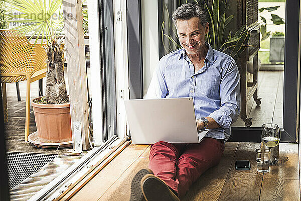 Porträt eines lächelnden Mannes  der am offenen Balkon einen Laptop benutzt