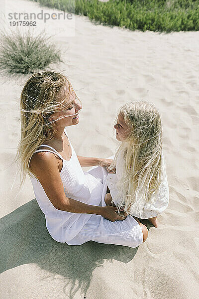 Lächelnde Mutter sitzt mit Tochter an einem sonnigen Tag am Strand