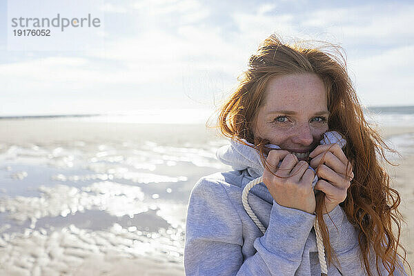 Redhead woman covering face in hooded shirt at beach