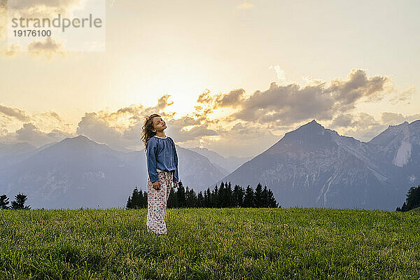 Mädchen steht bei Sonnenuntergang auf einer Wiese an den Bergen