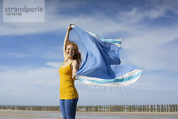 Glückliche Frau  die an einem sonnigen Tag mit einem Schal am Strand spielt
