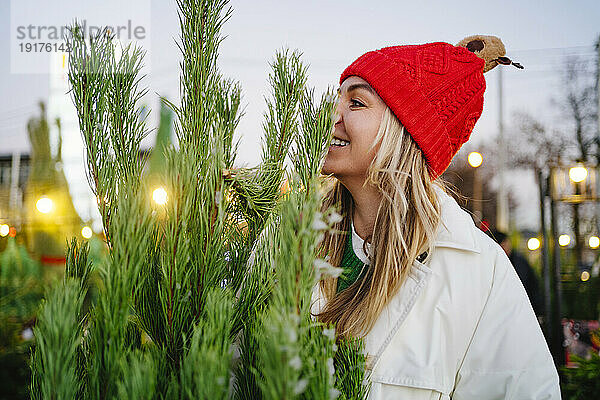 Smiling woman wearing knit hat near Christmas tree at market