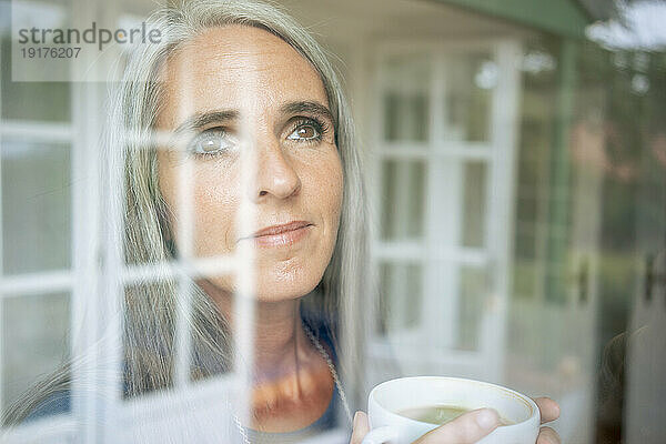 Reife Frau mit Teetasse  die durch das Fenster schaut