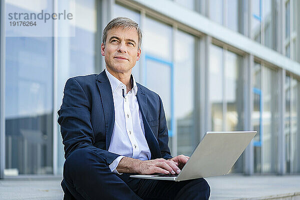 Geschäftsmann arbeitet am Laptop und sitzt vor dem Bürogebäude