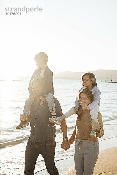 Eltern tragen Kinder auf Schultern und gehen am Strand entlang des Meeres