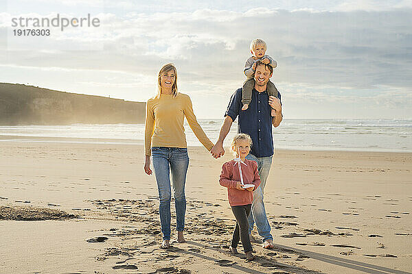 Glückliche Eltern halten Händchen und gehen mit Kindern am Strand spazieren