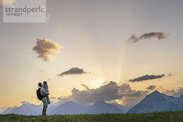 Father carrying daughter standing in meadow at sunset