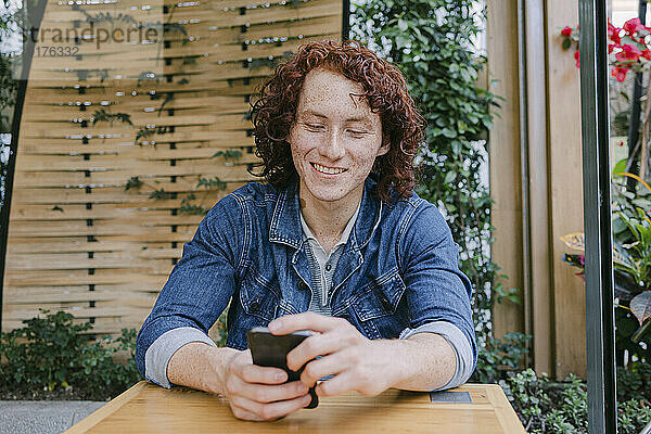 Glücklicher junger Mann  der sein Smartphone am Tisch im Café benutzt