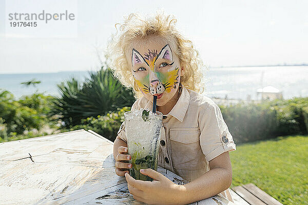 Mädchen mit Gesichtsbemalung trinkt Mojito im Garten an einem sonnigen Tag