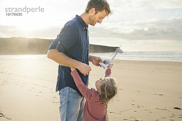 Glücklicher Vater und Tochter halten an einem sonnigen Tag ein Windturbinenmodell am Strand