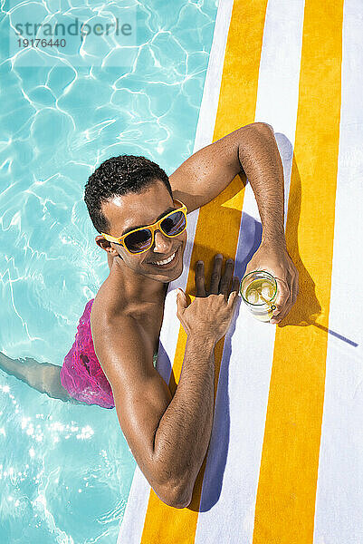 Lächelnder junger Mann mit Getränk  der sich am Pool auf ein Handtuch stützt