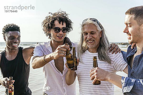 Lächelnde Freunde stoßen an einem sonnigen Tag am Strand auf Bierflaschen